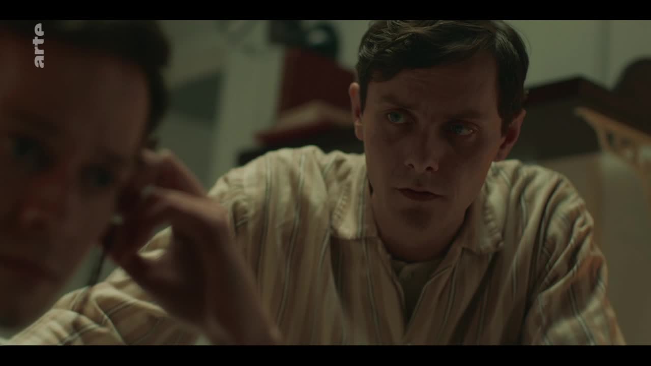 A man in a striped shirt leans forward, his gaze intense. Another person, out of focus, reaches a hand towards his face. A man in a striped shirt leans forward, his gaze intense. Another person, out of focus, reaches a hand towards his face.