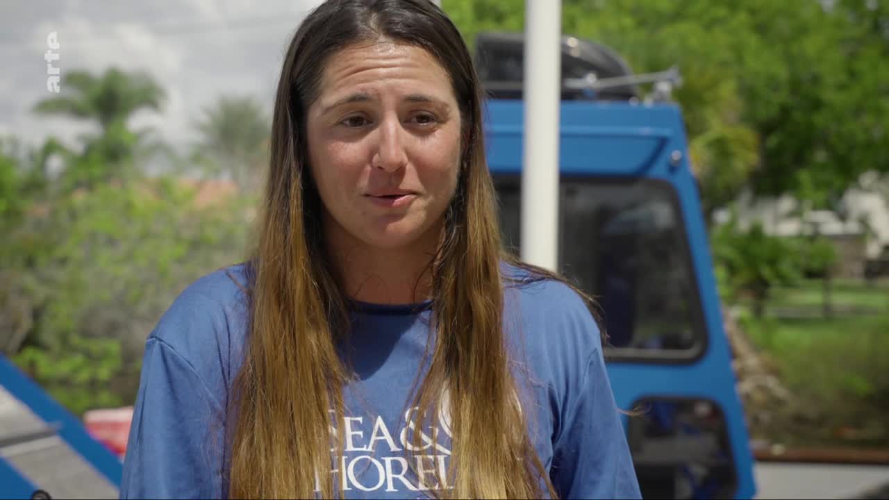 A woman in a blue t-shirt speaks, her long brown hair falling around her shoulders. Behind her, a bright blue vehicle, likely part of a research expedition from France, is parked under a bright sky. A woman in a blue t-shirt speaks, her long brown hair falling around her shoulders. Behind her, a bright blue vehicle, likely part of a research expedition from France, is parked under a bright sky.