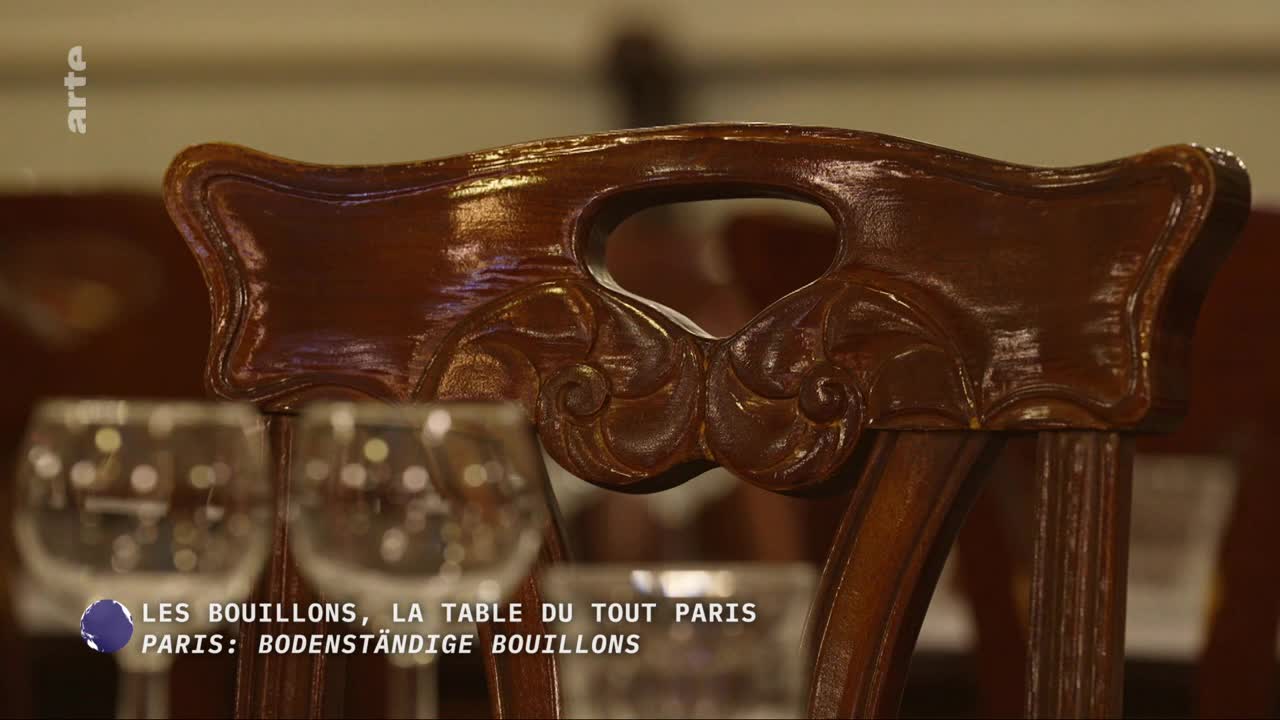 Two wine glasses sit on a table in front of an ornate wooden chair. The chair's back features a carved openwork design, hinting at a Parisian brasserie. Two wine glasses sit on a table in front of an ornate wooden chair. The chair's back features a carved openwork design, hinting at a Parisian brasserie.