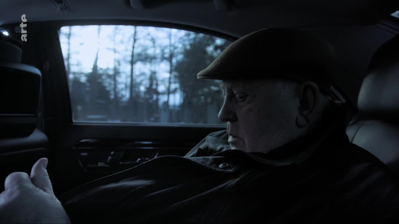 A man in a flat cap stares out the window of a car, the trees of France passing by. The soft glow from the Arte logo reflects on the screen in front of him.