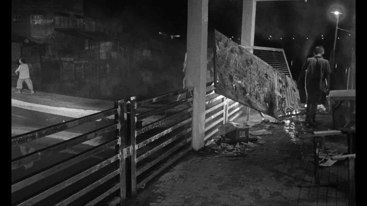 A young boy in a light shirt runs across a wet, dimly lit path. Further on, a man walks away from a large, tilted sign.
