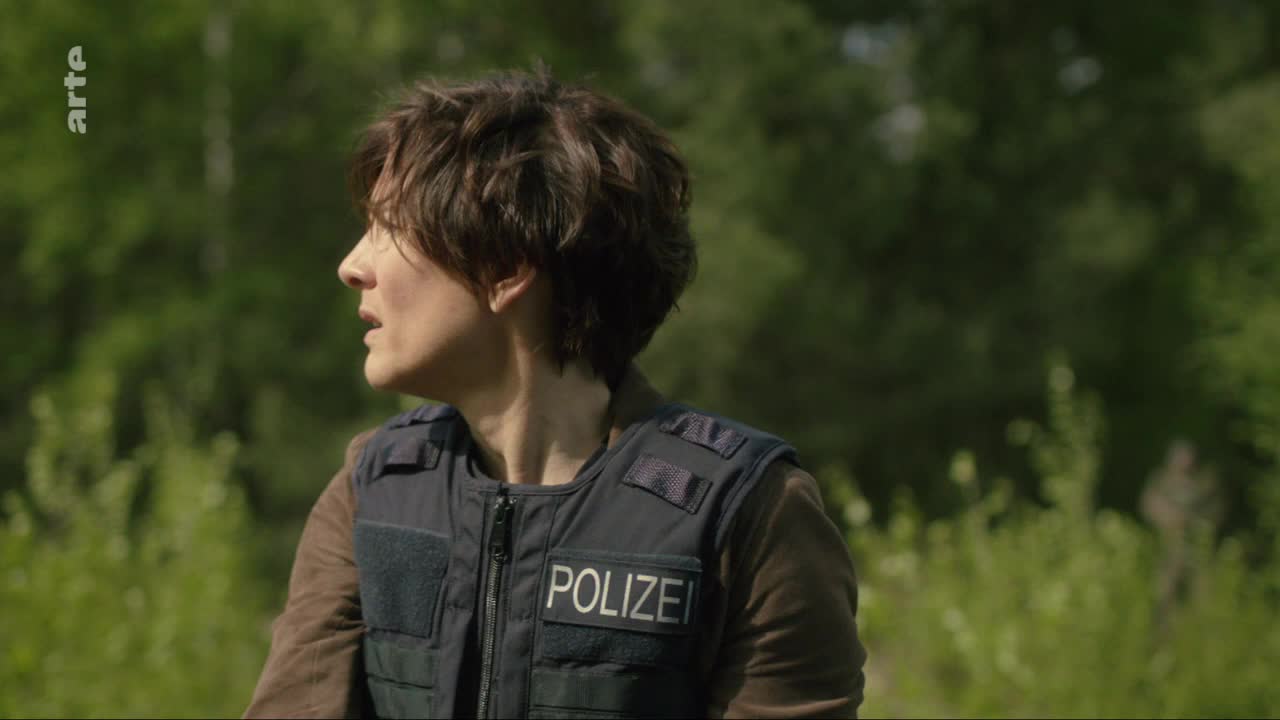 A woman in a tactical vest, marked "POLIZEI," scans the wooded area. Her gaze is fixed on something off-frame, her expression alert.