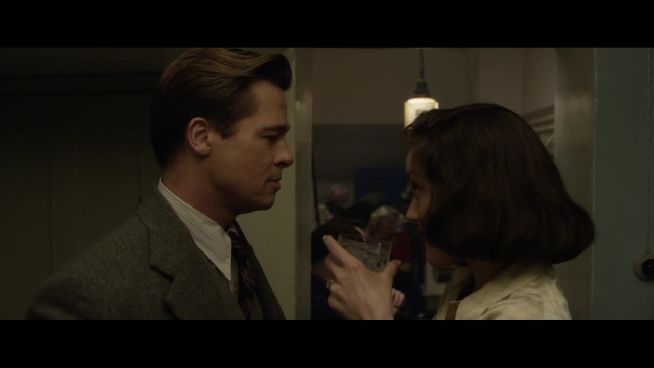 A man in a suit looks intently at a woman holding a drink. The muted lighting and the woman's posture suggest a quiet moment amidst a gathering.