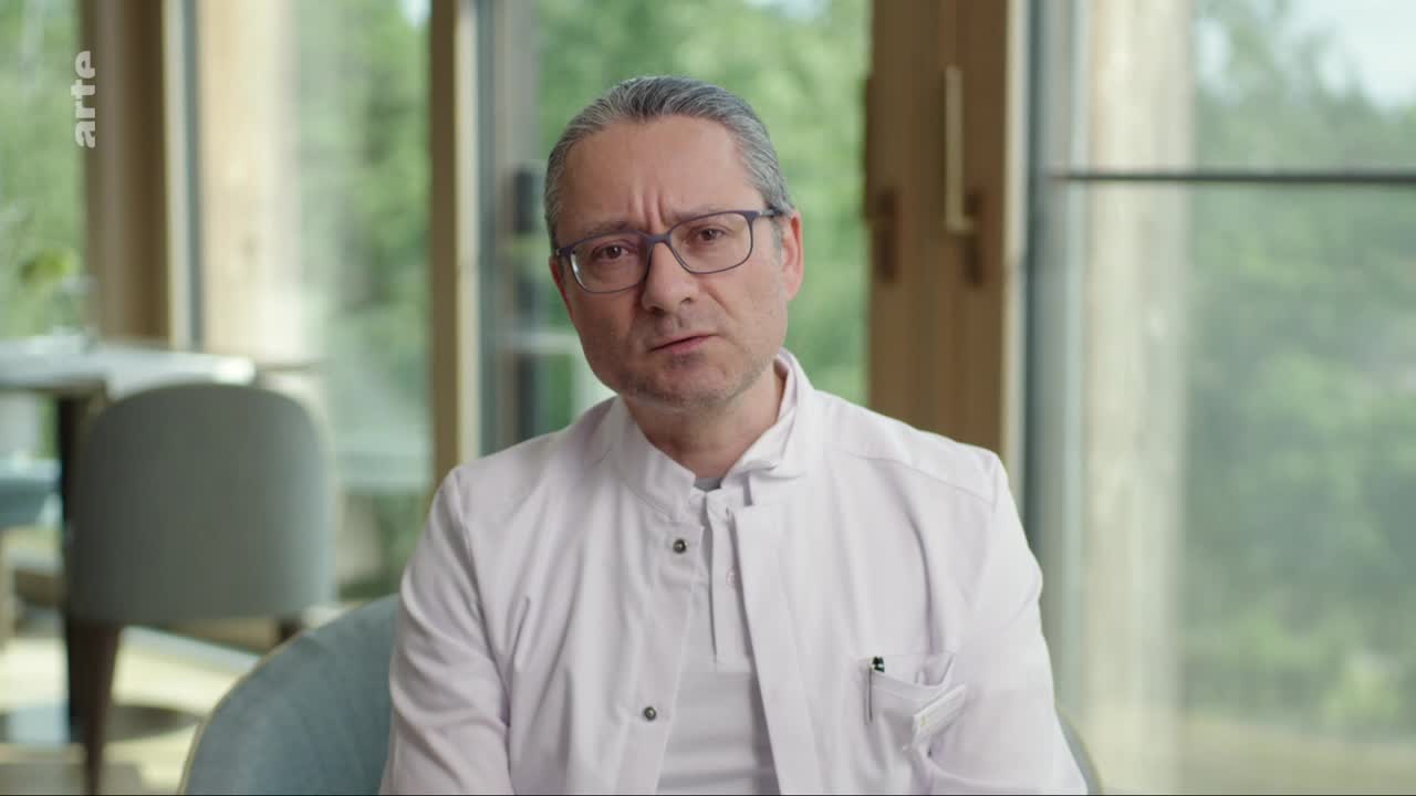 A man in a white lab coat speaks directly to the camera, his brow furrowed slightly. Behind him, a blurred background suggests a modern interior with large windows.