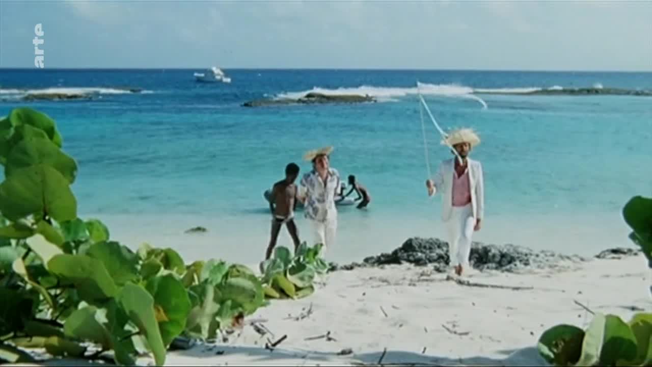 Two men in straw hats walk along a white sand beach toward the turquoise sea. One man, dressed in a white suit and pink shirt, carries a fishing rod.