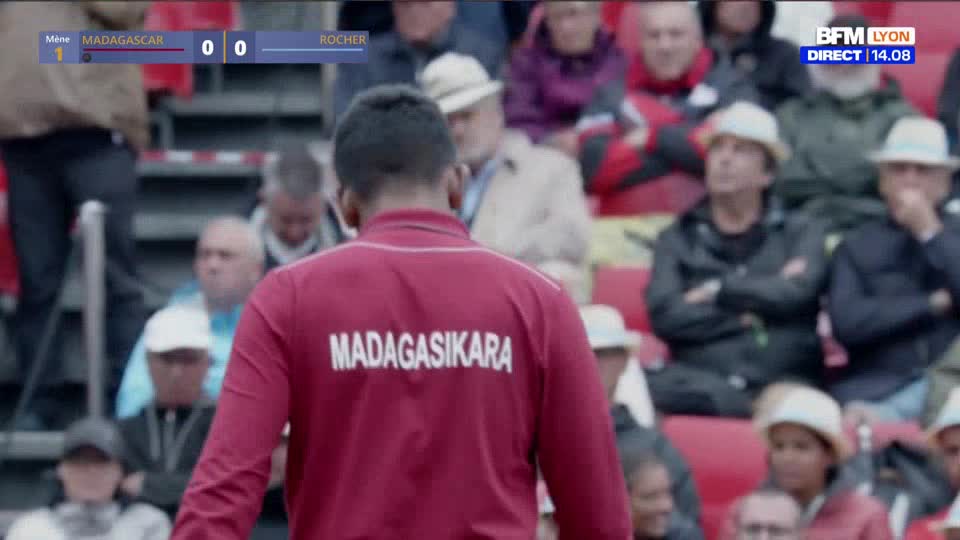The player in the red jacket, "Madagasikara" emblazoned on the back, stands facing the field. Spectators in hats and jackets watch from the stands, the BFM Lyon broadcast logo visible in the corner.
