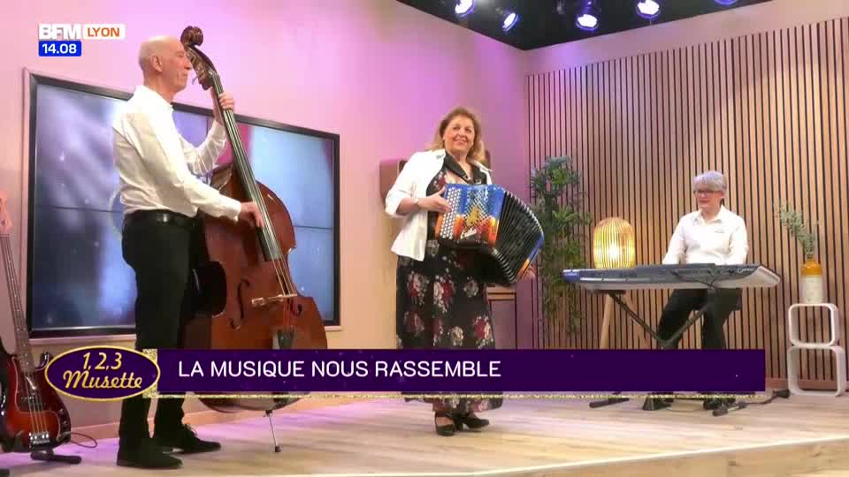 On the BFM Lyon set, a trio is playing. The woman in the middle, smiling, pumps her accordion, while the man on the left bows his upright bass.
