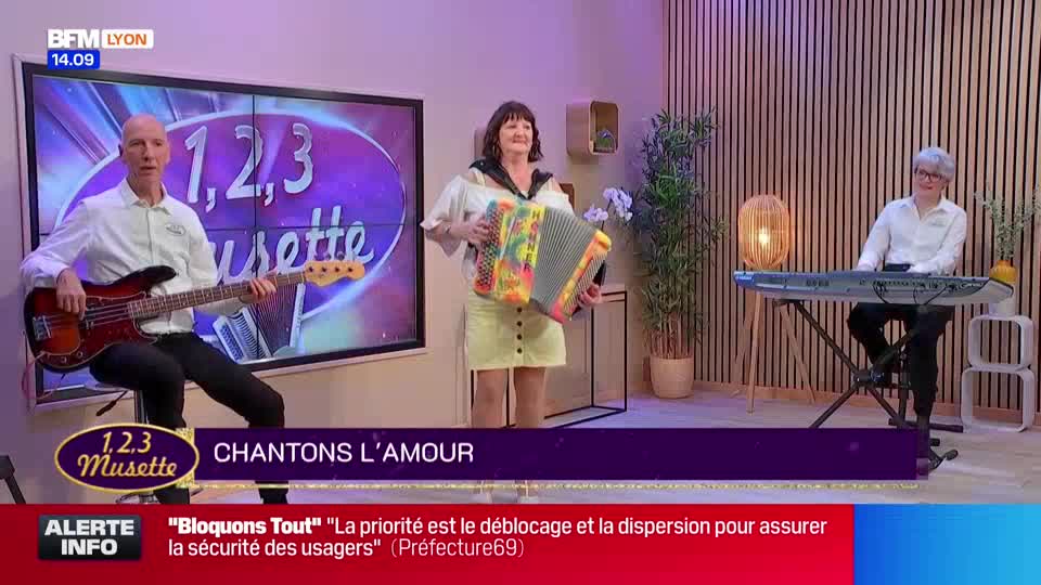 On a BFM Lyon set, a trio performs: a bassist, an accordion player, and a keyboardist. The accordionist, in a yellow skirt, smiles as she plays.
