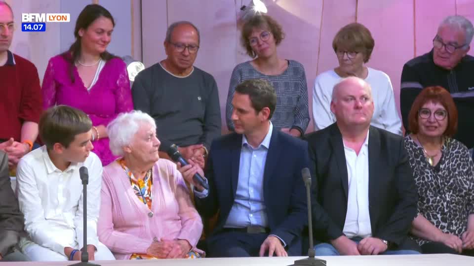 A man in a suit holds a microphone, speaking to an elderly woman in a pink sweater. Behind them, a group of people watch, some with slight smiles.
