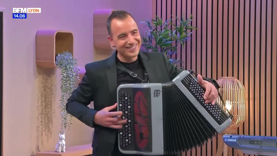 A man in a dark suit smiles as he plays the accordion. The BFM Lyon logo is visible in the corner, and a keyboard sits nearby.
