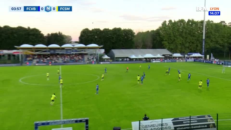 Players in yellow and blue jerseys are spread across the pitch, the ball is in play. The scoreboard on BFM Lyon shows the score is still nil-nil, early in the match.
