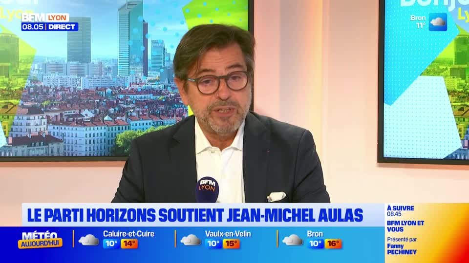 A man in glasses, wearing a dark suit jacket, is speaking into a microphone with the BFM Lyon logo. Behind him, a cityscape of Lyon is visible on a screen, with weather information displayed below.
