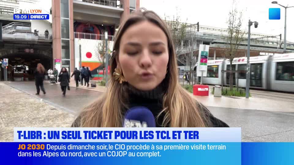 A reporter from BFM Lyon is speaking into a microphone, with a tram gliding past behind her. People walk on the pedestrian area, and the rain-slicked tracks reflect the overcast sky.
