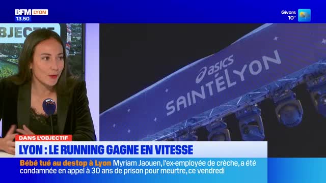 A woman speaks at a BFM Lyon desk, while a large blue banner with "SAINTÉLYON" and the Asics logo hangs above, illuminated by spotlights. The banner appears to be part of a race event in France. A woman speaks at a BFM Lyon desk, while a large blue banner with "SAINTÉLYON" and the Asics logo hangs above, illuminated by spotlights. The banner appears to be part of a race event in France.