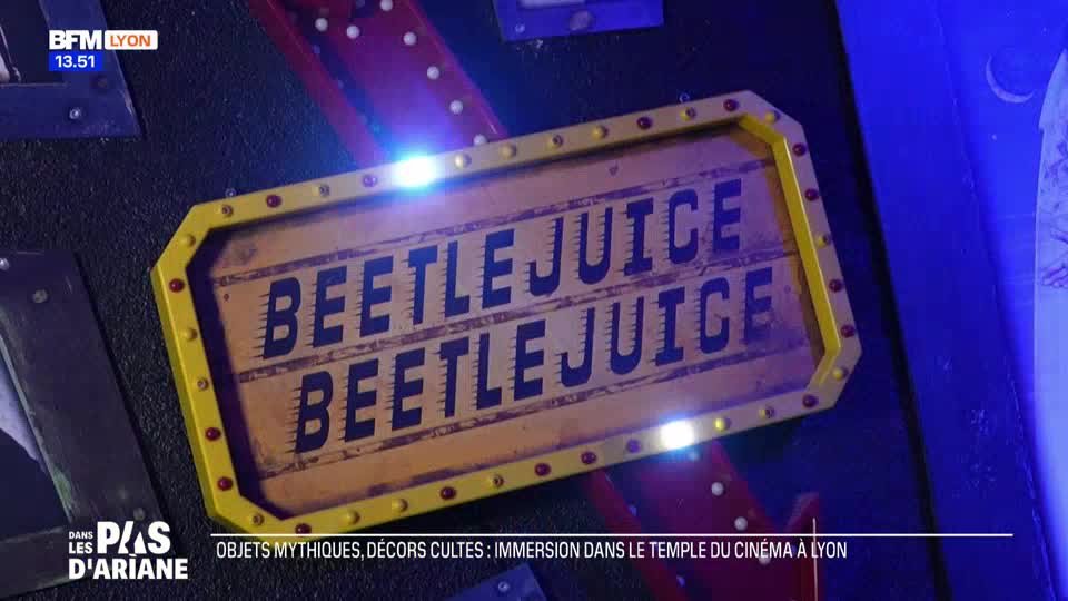 A weathered, yellow sign with red bolts and two lines of bold, black text reading "BEETLEJUICE" hangs on a dark wall. The sign is illuminated by bright, blue-white lights, hinting at a theatrical or exhibition space in Lyon, France. A weathered, yellow sign with red bolts and two lines of bold, black text reading "BEETLEJUICE" hangs on a dark wall. The sign is illuminated by bright, blue-white lights, hinting at a theatrical or exhibition space in Lyon, France.
