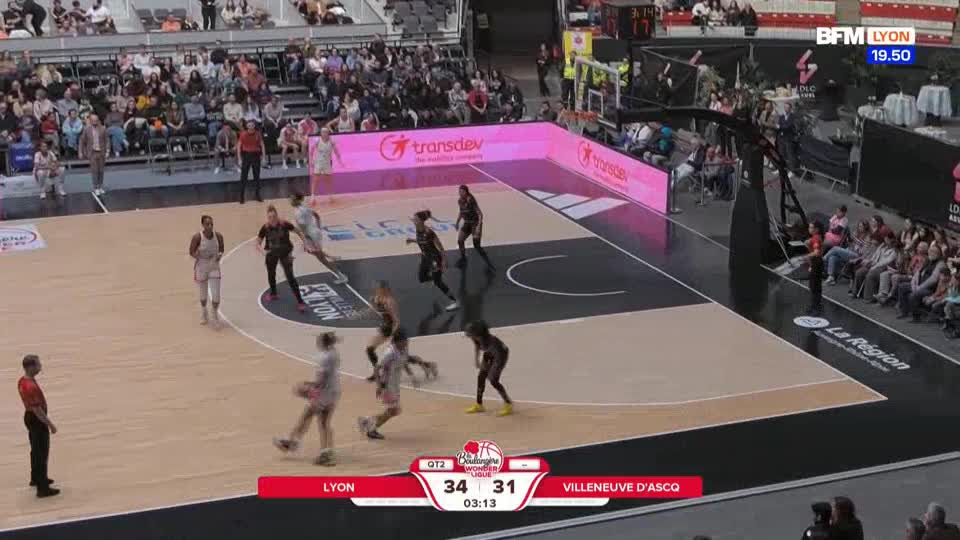 A player in a white uniform drives towards the basket, while another in black defends. The scoreboard shows Lyon leading Villeneuve d'Asco 34 to 31. A player in a white uniform drives towards the basket, while another in black defends. The scoreboard shows Lyon leading Villeneuve d'Asco 34 to 31.