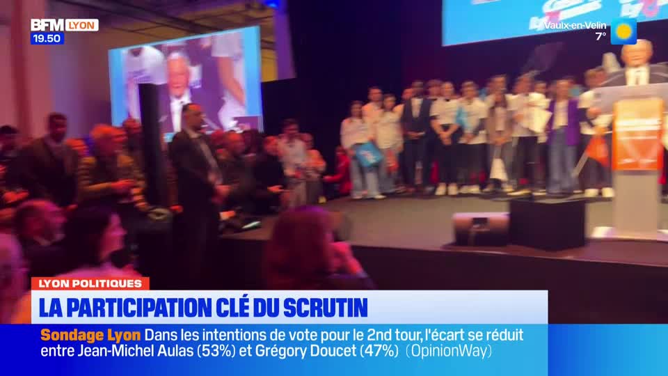 A group of people stands on a stage in front of a large screen displaying a man's face. Below, a news ticker from BFM Lyon announces polling data for the second round of an election.