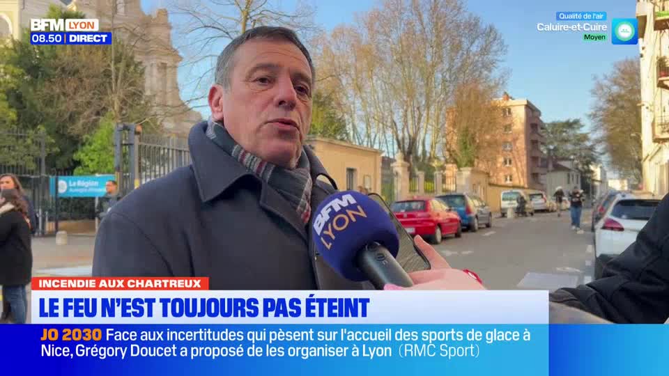 A man in a dark coat and scarf speaks into a BFM Lyon microphone. Cars are parked along the street behind him, and a few people walk in the distance.