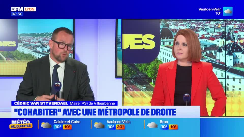 A man in a suit and tie speaks into a microphone, his glasses reflecting the studio lights. Beside him, a woman in a bright red blazer listens intently, her red hair framing her face.