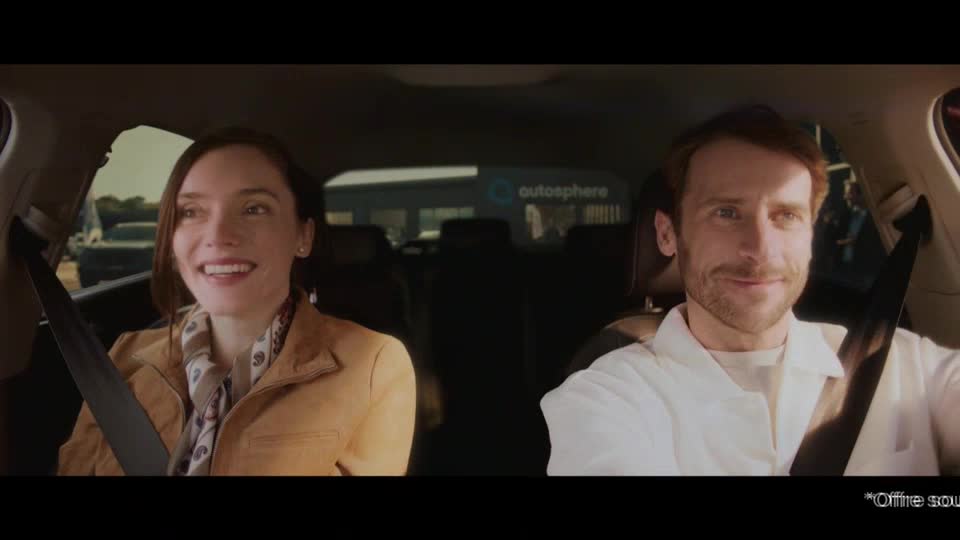 The car is moving past a building with "autosphere" written on it. The woman in the passenger seat laughs, her eyes bright. The car is moving past a building with "autosphere" written on it. The woman in the passenger seat laughs, her eyes bright.