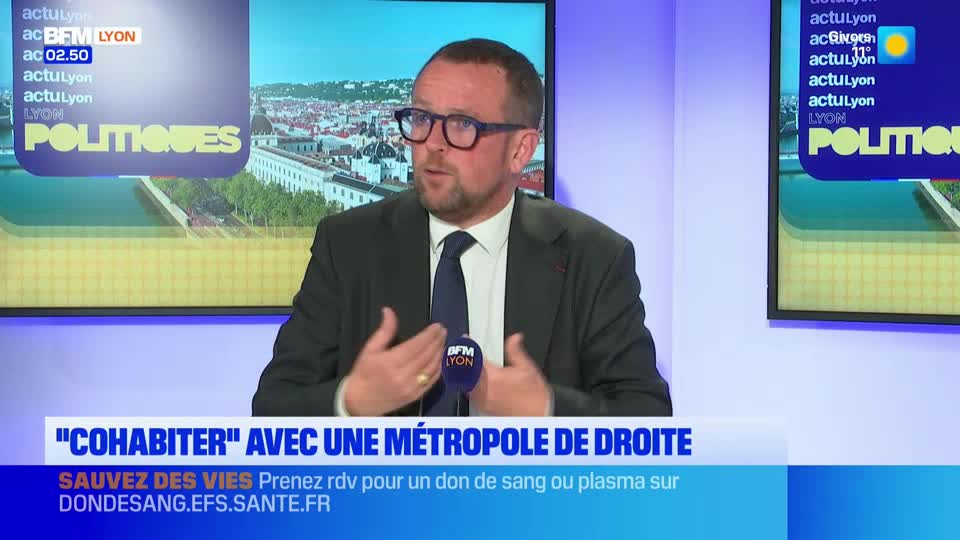 A man in a suit and tie gestures with his hands while speaking into a microphone. A screen behind him displays graphics for BFM Lyon's "Politiques" segment. A man in a suit and tie gestures with his hands while speaking into a microphone. A screen behind him displays graphics for BFM Lyon's "Politiques" segment.