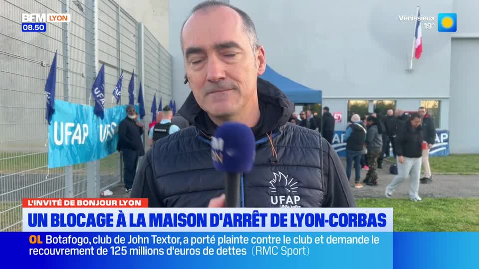 A reporter stands with a microphone in front of a crowd gathered outside a building in France. Blue flags bearing the UFAP logo flutter in the breeze, and a large banner with the same insignia hangs on a fence. A reporter stands with a microphone in front of a crowd gathered outside a building in France. Blue flags bearing the UFAP logo flutter in the breeze, and a large banner with the same insignia hangs on a fence.