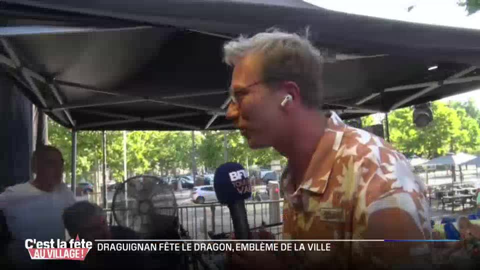 A reporter for BFM Lyon holds a microphone, speaking into it as a large fan whirs behind him. The scene appears to be an outdoor festival in Draguignan, celebrating their city's emblem.