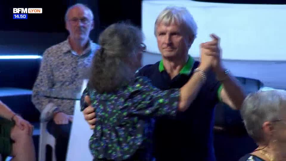 A couple dances in front of a blue backdrop, their hands clasped. A man in a patterned shirt watches from the background, seated.