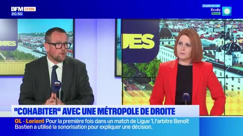 A man in a suit and glasses speaks into a microphone, while a woman in a red blazer listens intently. Behind them, a screen displays a cityscape and the BFM Lyon logo.