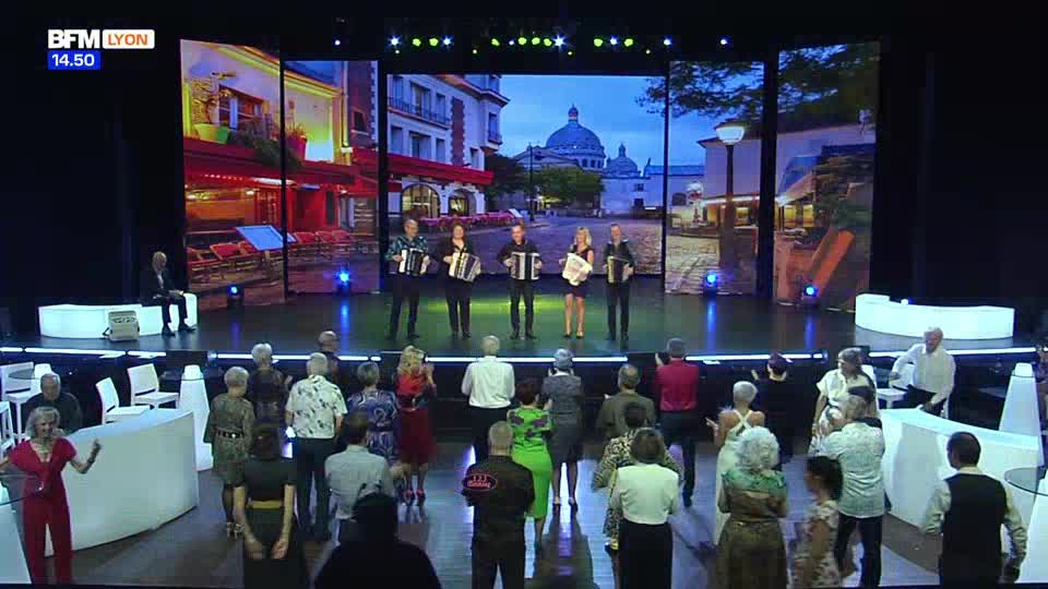 Five musicians stand on a stage, each with an accordion, as a BFM Lyon broadcast plays behind them. A crowd watches from the audience, some standing and some seated.