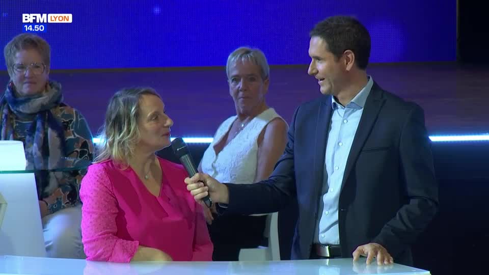 A man in a dark suit interviews a woman in a bright pink top, holding a microphone to her. Behind them, two other women watch the exchange.