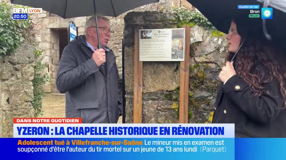 A man in a grey jacket holds an umbrella, speaking near a stone wall. A woman with dark curly hair, also under an umbrella, stands to his right.