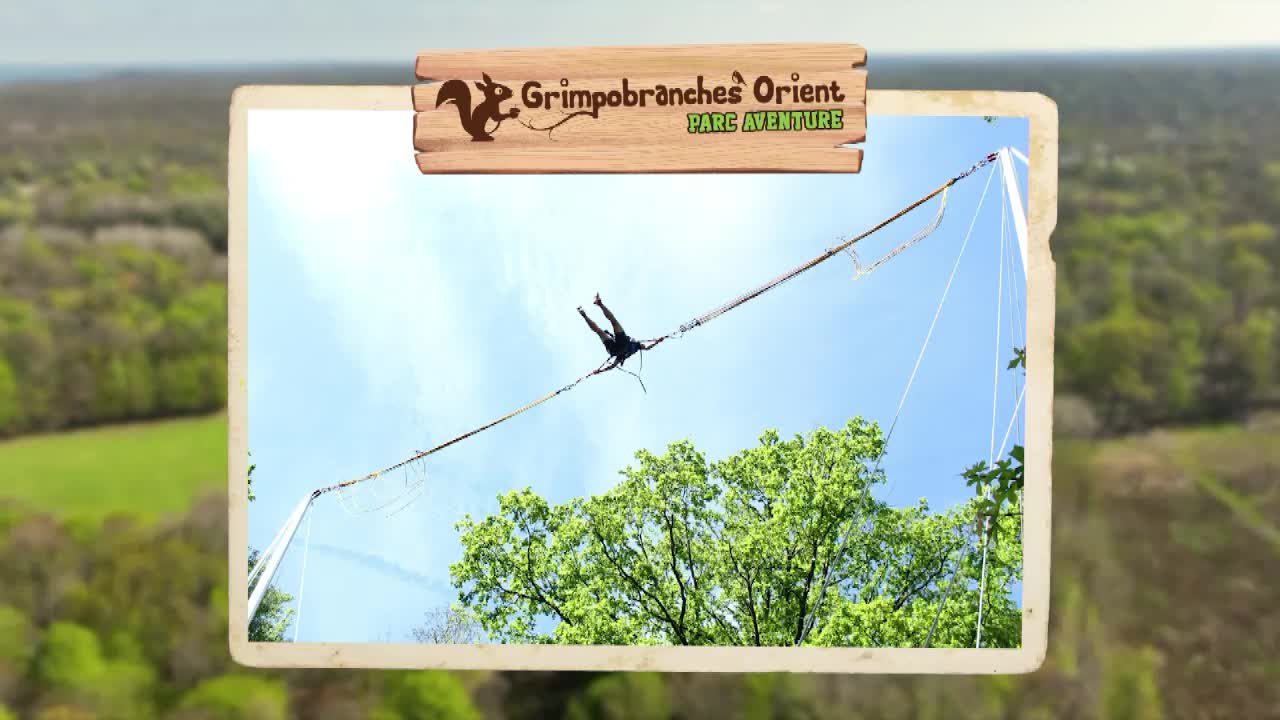 A person is suspended mid-air, dangling from a cable at GrimpoBranches Orient Parc Aventure in France, as seen on Canal 32. They're silhouetted against the bright sky, with the green treetops below.
