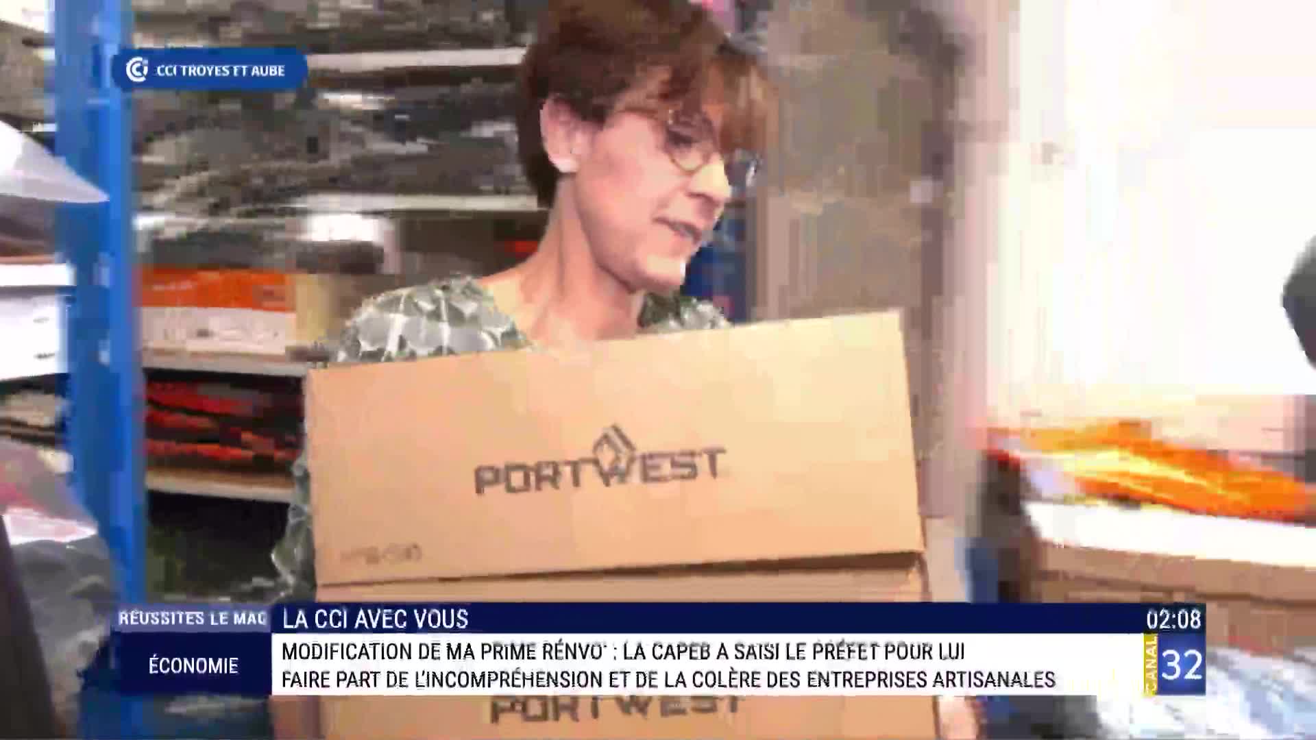 A person in France, on Canal 32, carries a stack of cardboard boxes labeled "Portwest" towards the camera. The boxes obscure their lower body as they move through a cluttered storage area.
