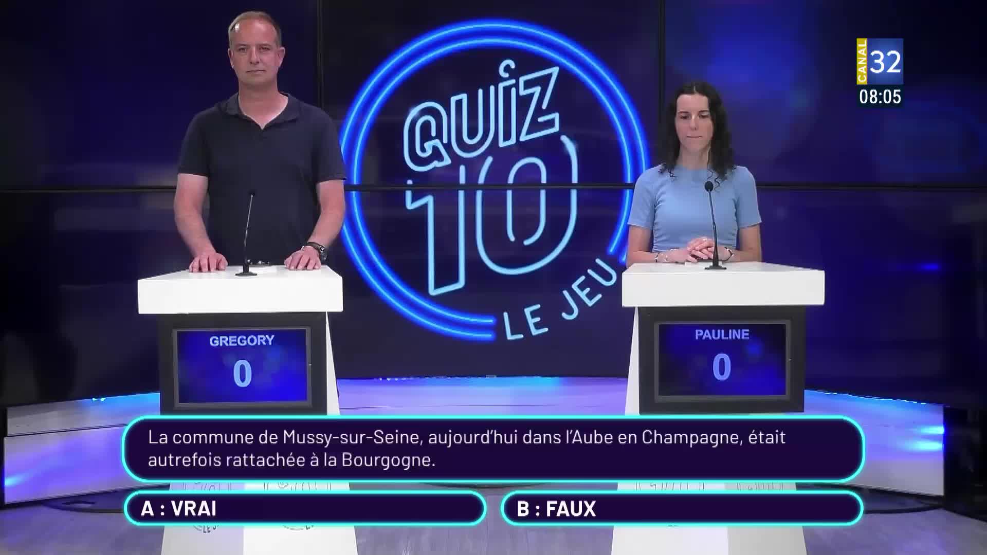 Two contestants stand at podiums, facing forward on the set of a French game show, Canal 32. A question about a town in France is displayed, and the two are ready to answer.
