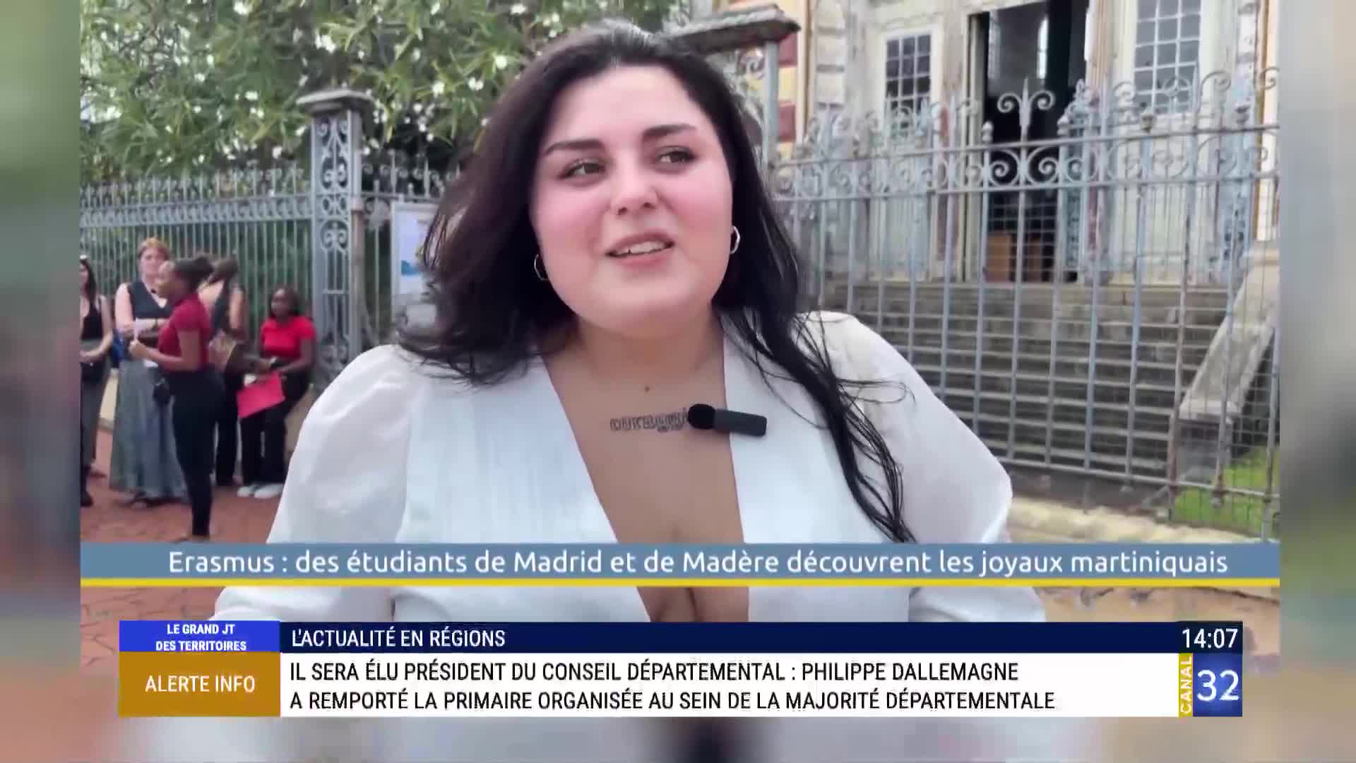 A woman in a white blouse speaks directly to the camera, a microphone clipped to her neckline. Behind her, a group of people stand near a wrought-iron gate, likely part of a segment on Canal 32 from France.
