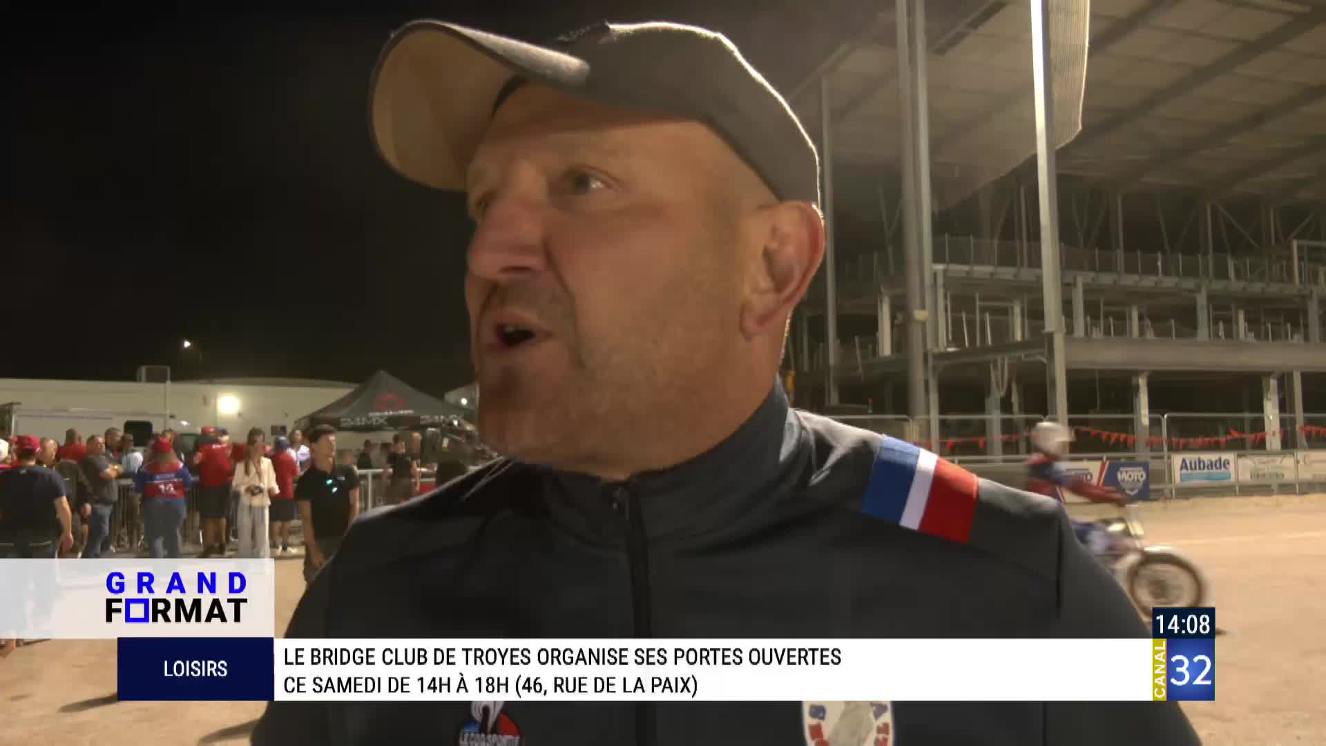 A man in a cap is speaking, likely for Canal 32, with a crowd of people behind him. A motorcycle racer is visible in the background, and the event is taking place in France.
