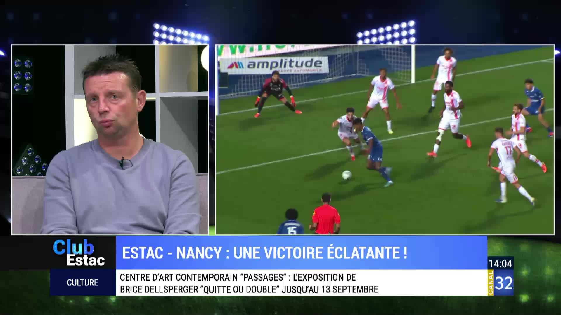 On Canal 32, a man in a grey shirt looks at the camera while a football game plays on the screen behind him. The blue-clad player dribbles the ball toward the goal, pursued by several players in white.
