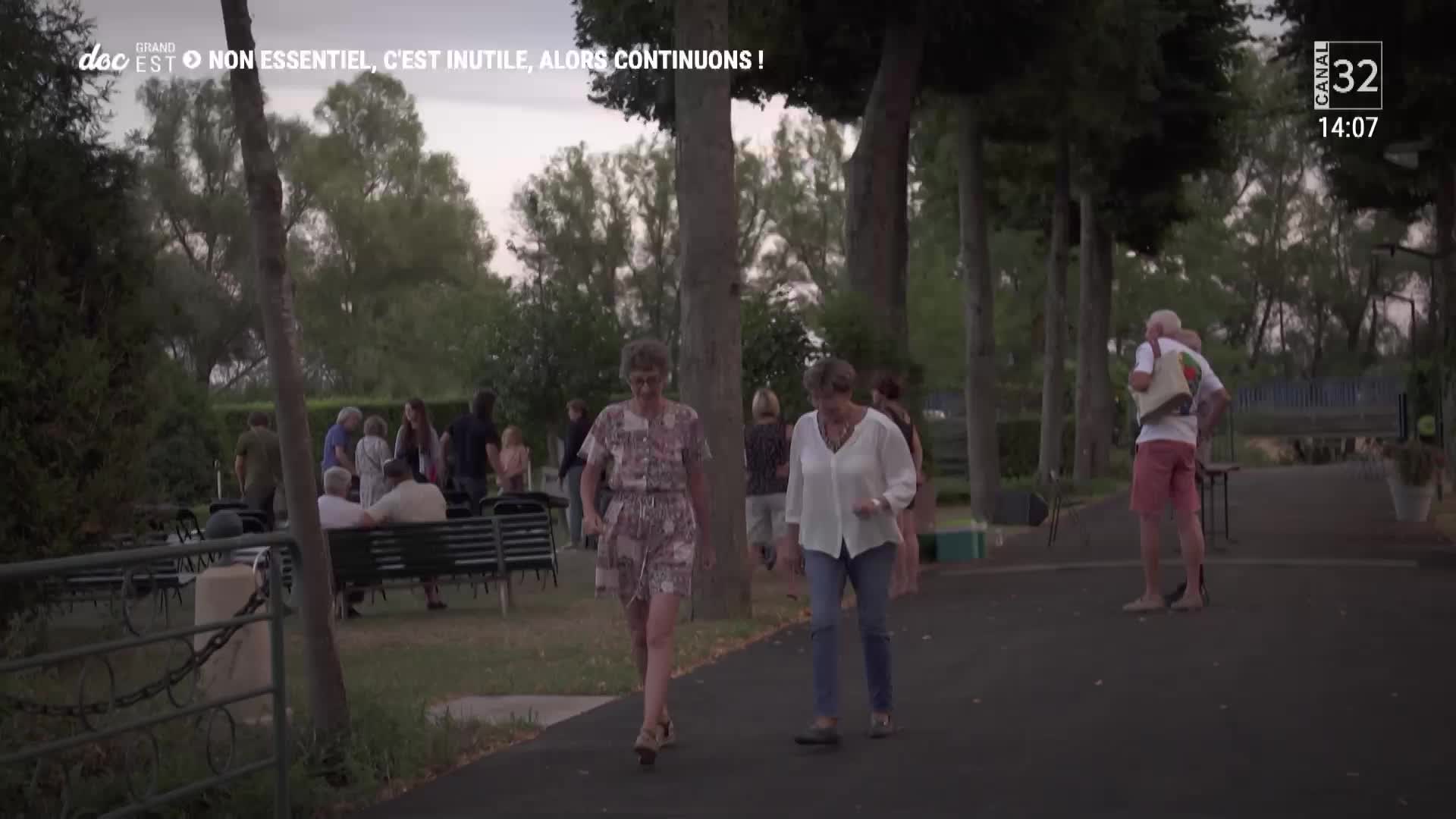 Two women stroll down a paved path, past a group of people gathered in a park. The Canal 32 logo and time appear in the upper right corner, suggesting this is French television.

