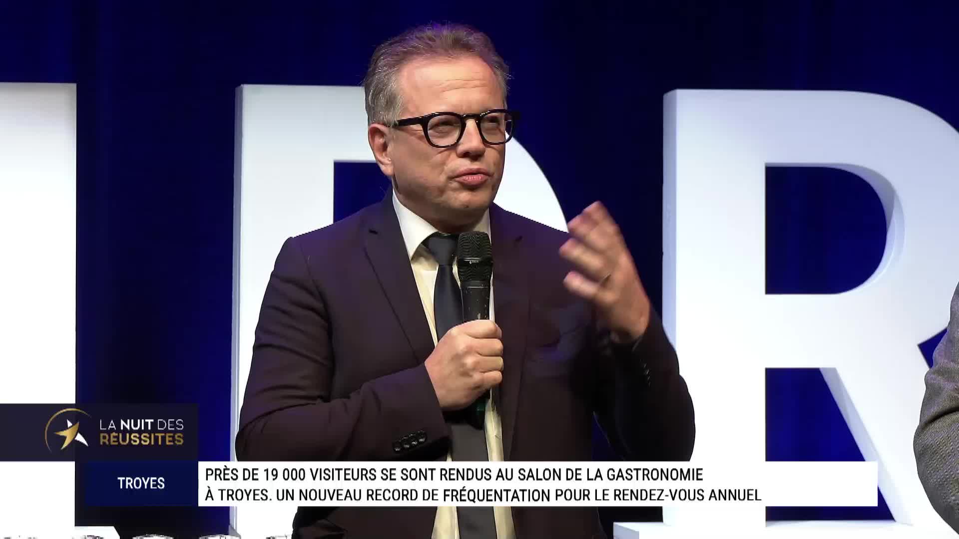 A man in a suit, wearing glasses, speaks into a microphone, gesturing with his right hand. The backdrop features a large white "R" against a dark blue background, likely for a broadcast on Canal 32 in France.
