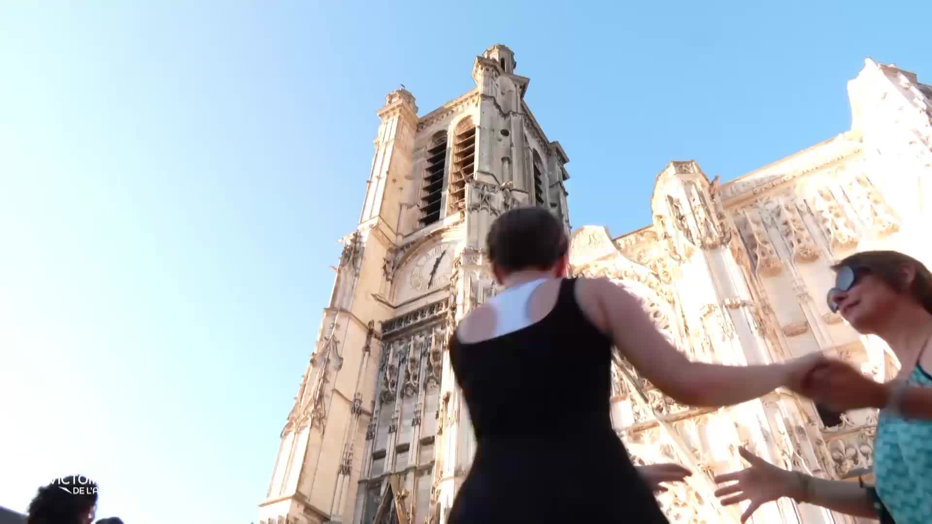 Two women dance in front of a grand church, their arms outstretched as they move. The camera angle looks up at the building, with the blue sky overhead.
