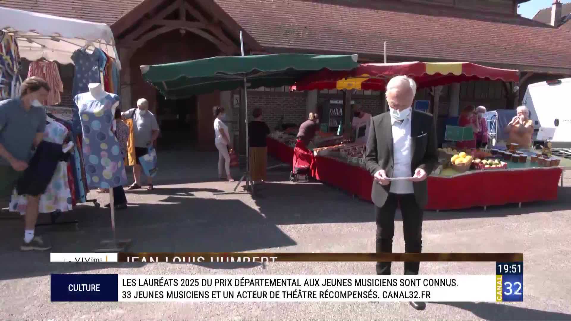 A man in a suit stands by a market stall, holding a piece of paper. People mill about behind him, browsing goods under colorful awnings. 
