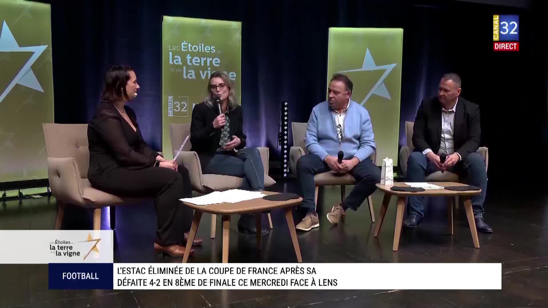 A woman with glasses speaks into a microphone, gesturing with her hands. Beside her, a man in a blue cardigan listens intently.