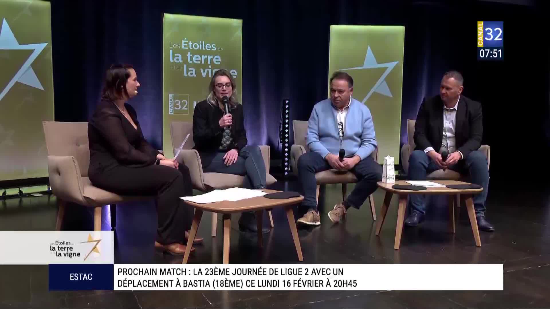 A woman with glasses speaks into a microphone, seated on a chair. Beside her, a man in a light blue cardigan leans forward, holding his own microphone.