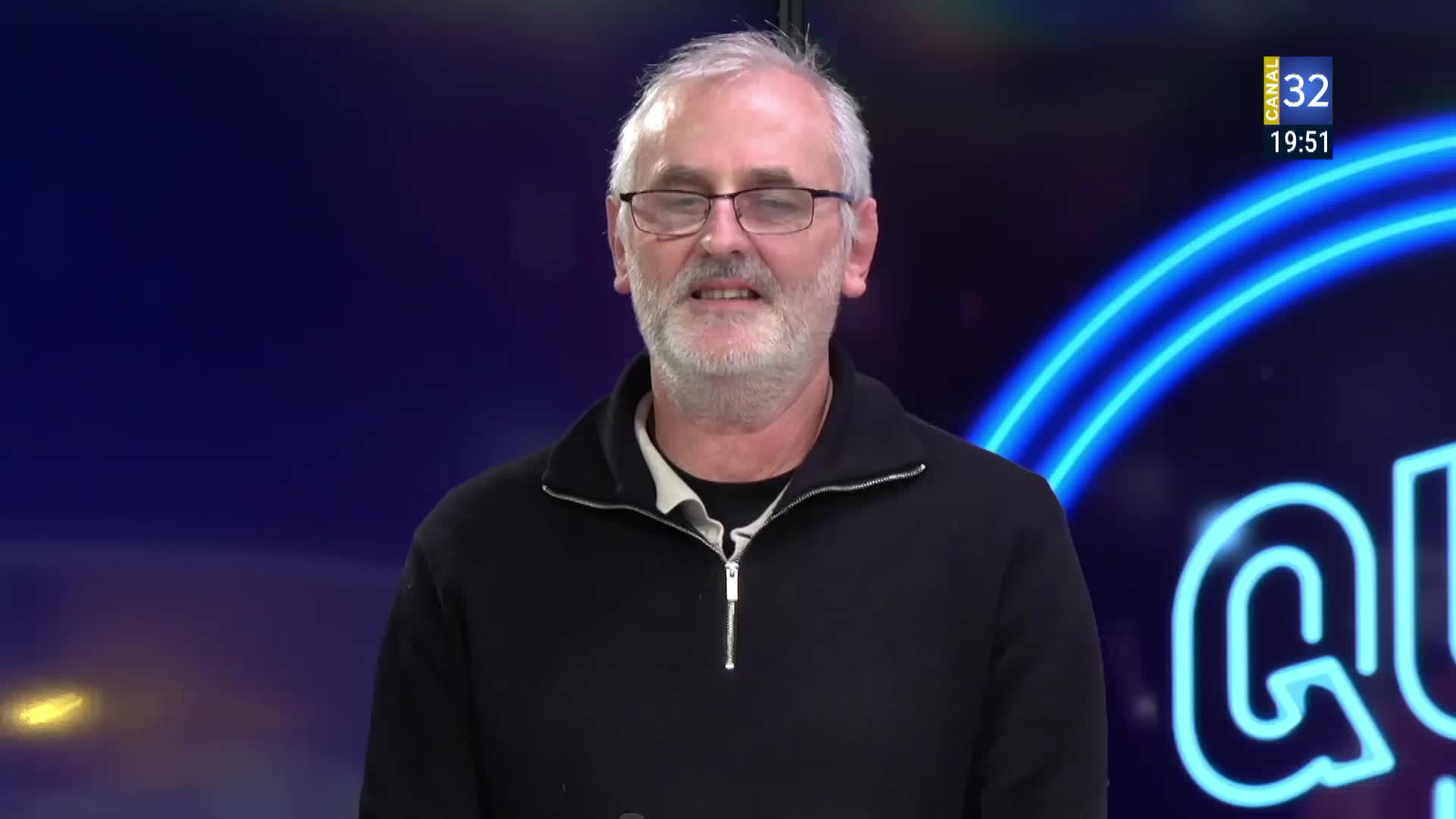 A man with glasses and a gray beard stands before a dark blue background with neon lights. A logo for Canal 32 appears in the upper right corner.