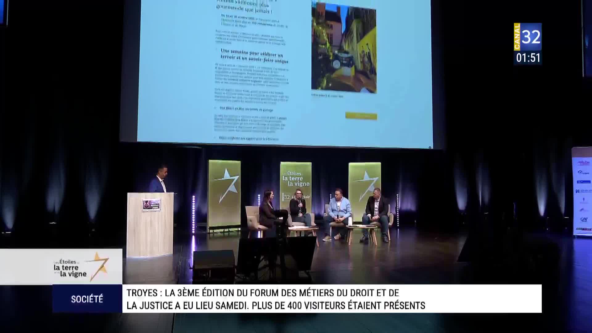 A man stands at a podium on a stage in front of a large screen displaying text and an image. Four people sit on chairs in a semicircle on the stage, engaged in discussion.