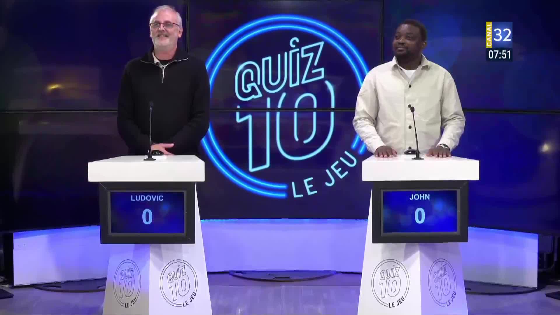 Two men stand at their podiums on the Canal 32 quiz show, "Quiz 10." The scoreboard for each contestant, Ludovic and John, displays a score of zero.