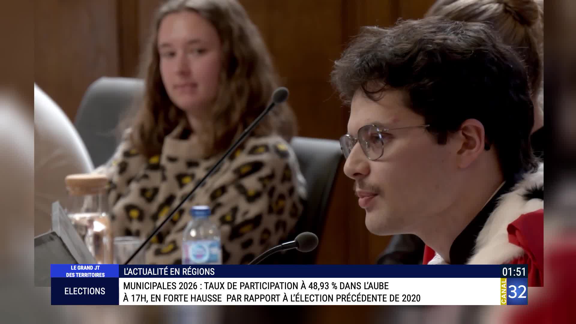A young man with glasses and a mustache speaks into a microphone, wearing a robe with fur trim. Behind him, a woman with long brown hair looks towards the camera. A graphic from Canal 32 displays election results for "Municipalités 2026." A young man with glasses and a mustache speaks into a microphone, wearing a robe with fur trim. Behind him, a woman with long brown hair looks towards the camera. A graphic from Canal 32 displays election results for "Municipalités 2026."