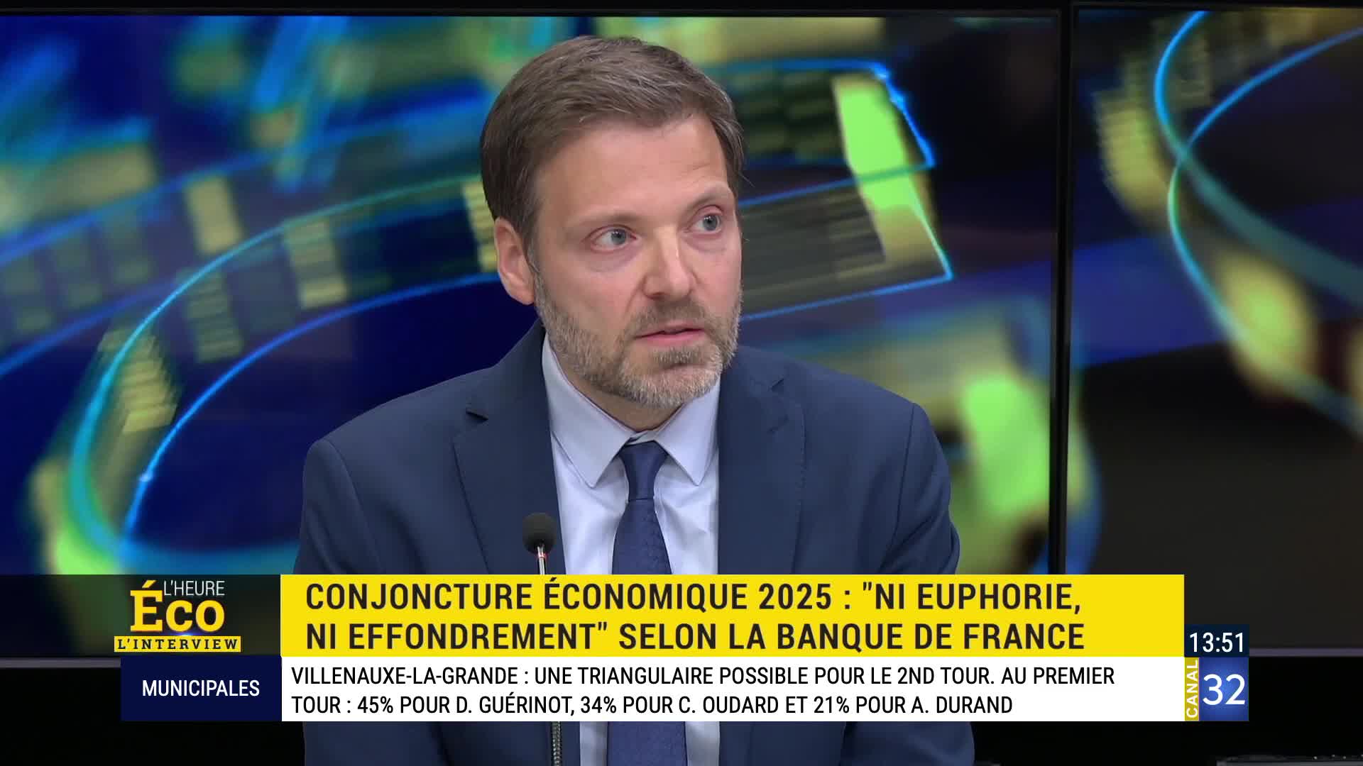 A man in a suit speaks into a microphone, his expression serious. Behind him, a screen displays graphics and text from Canal 32, including a headline about the French economy. A man in a suit speaks into a microphone, his expression serious. Behind him, a screen displays graphics and text from Canal 32, including a headline about the French economy.