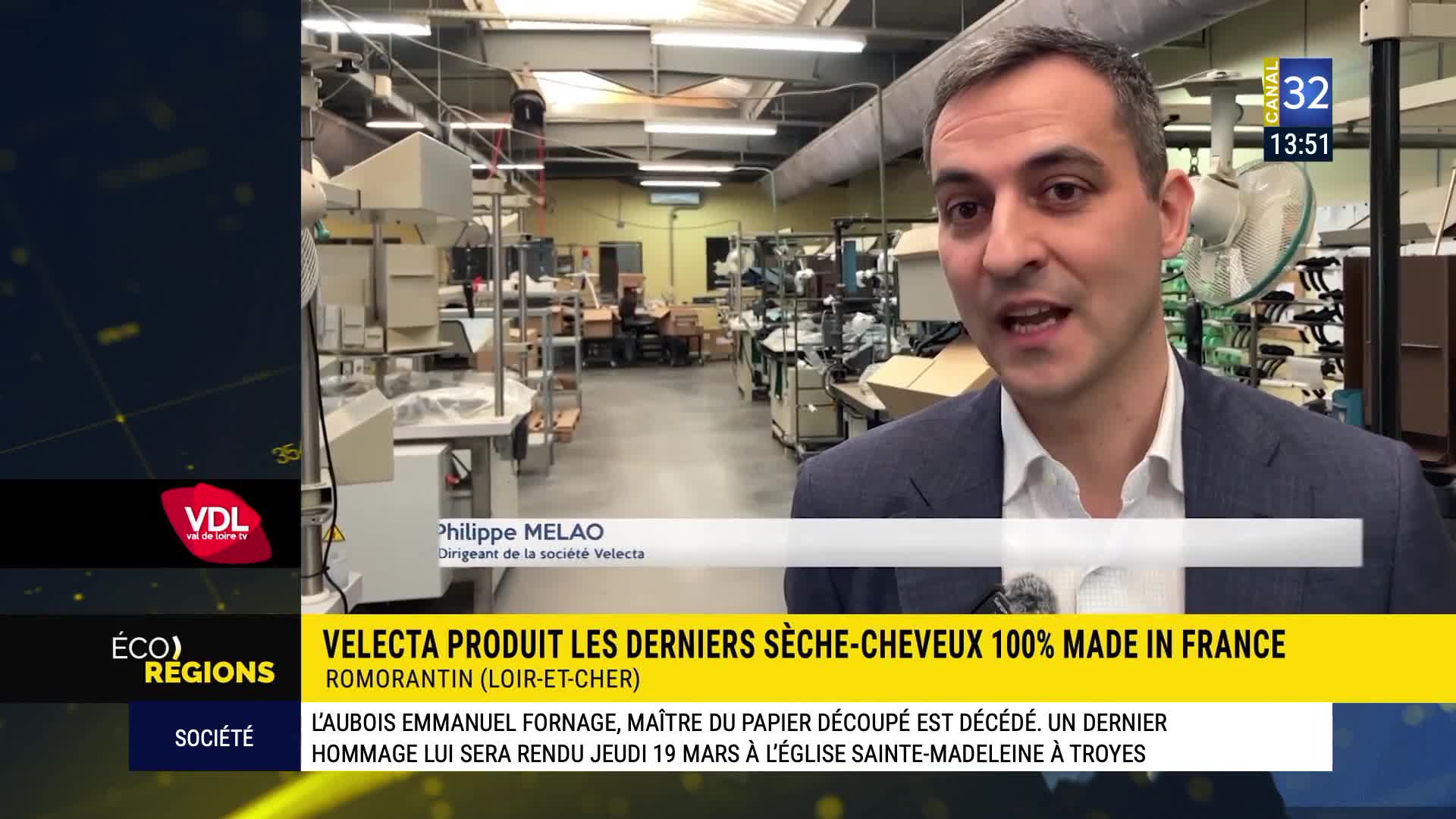 A man in a suit speaks directly to the camera from inside a factory in France. Behind him, rows of machinery and workers are visible, suggesting a busy production environment. A man in a suit speaks directly to the camera from inside a factory in France. Behind him, rows of machinery and workers are visible, suggesting a busy production environment.